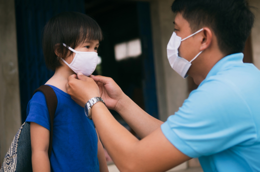  Asian Father Wearing Protective Mask Puts Protective Cloth Face Mask On His Little Child, Schoolkid With Backpack Is Ready To